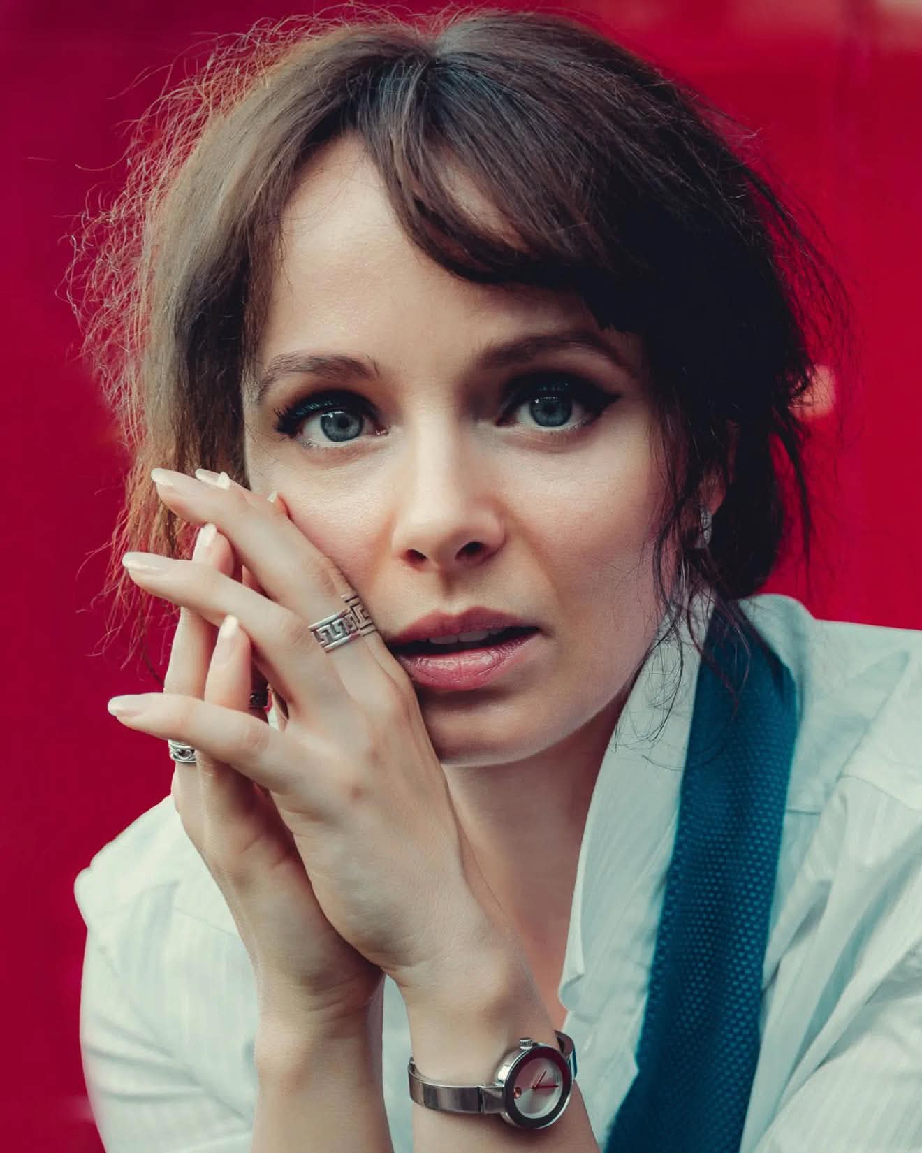 Olya Aman against a red wall, fingertips pressed together at her cheek